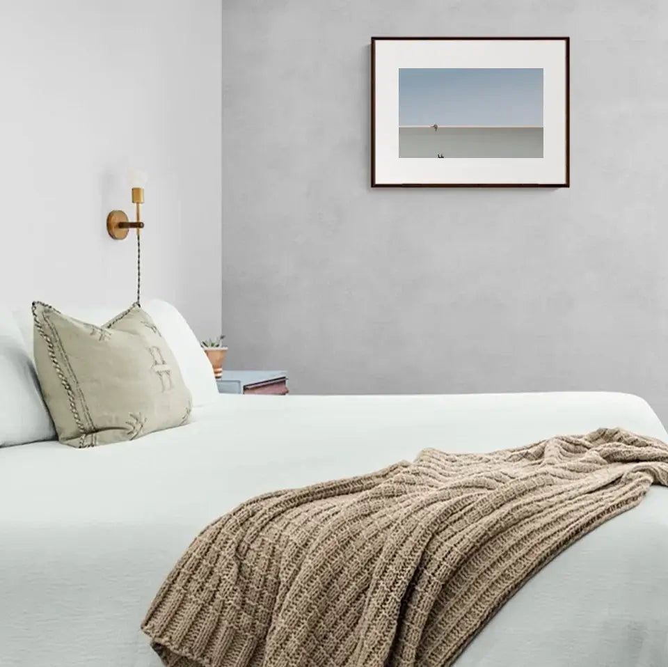 Minimalist bedroom with cozy blanket and framed beach photograph on gray wall.