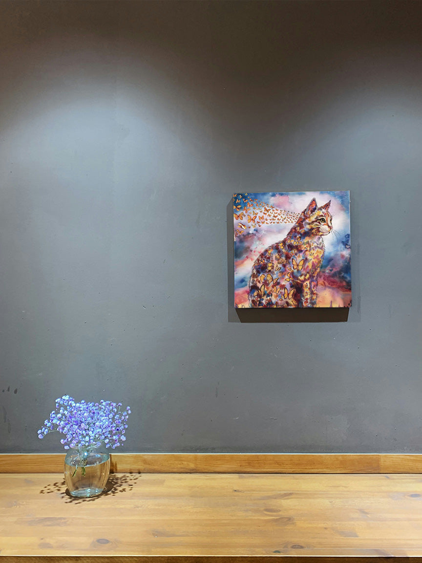 “Wings of Life” - A captivating painting of a cat adorned with butterflies hangs on a grey wall, complemented by a vase of purple flowers below.